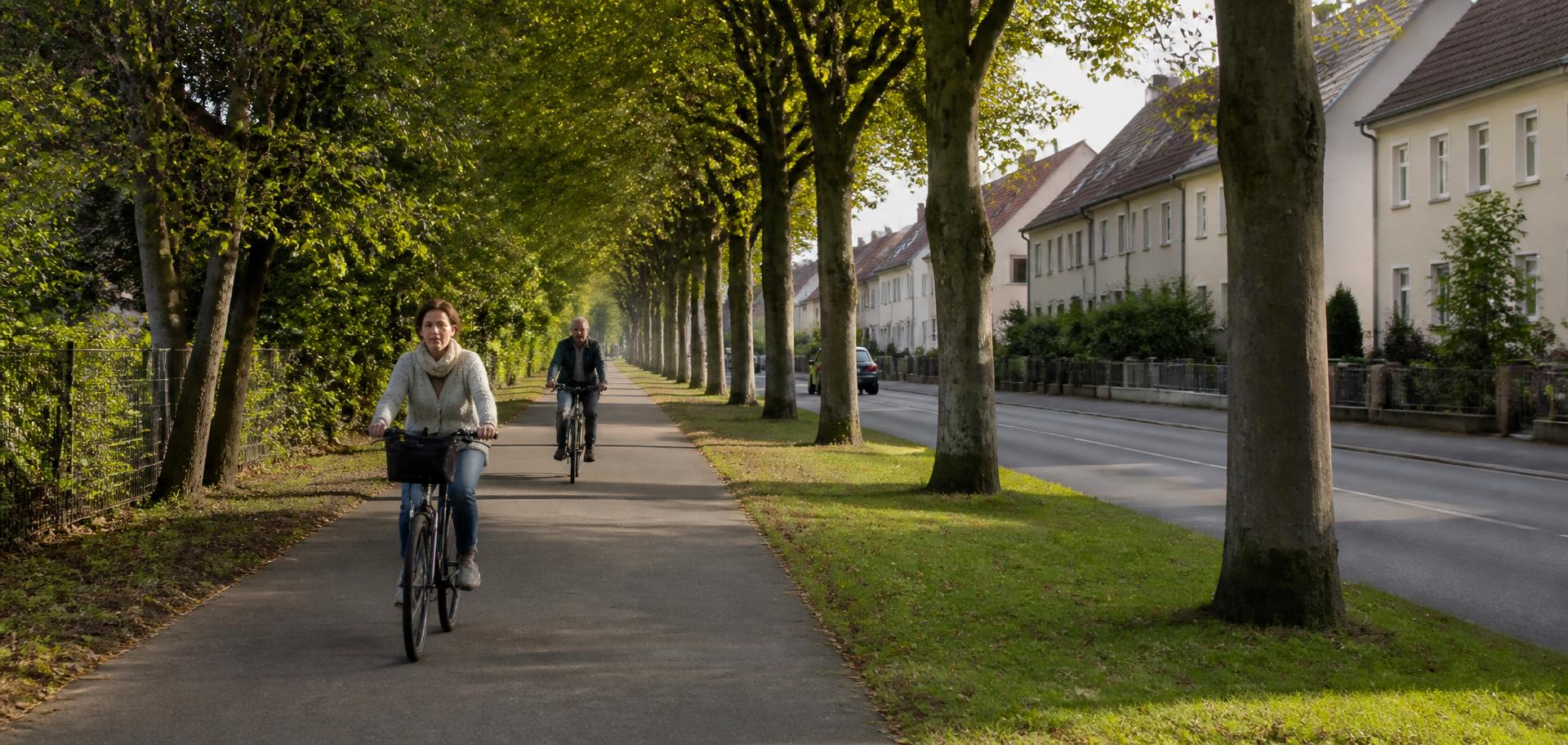 Zwei Radfahrende auf einem baumbestandenen Radweg entlang einer Wohnstraße